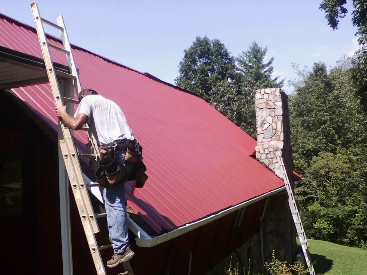 Roofer on a ladder beside a red metal roof and stone chimney — Soffit & Fascia Repair by Roofing Brothers in Waconia